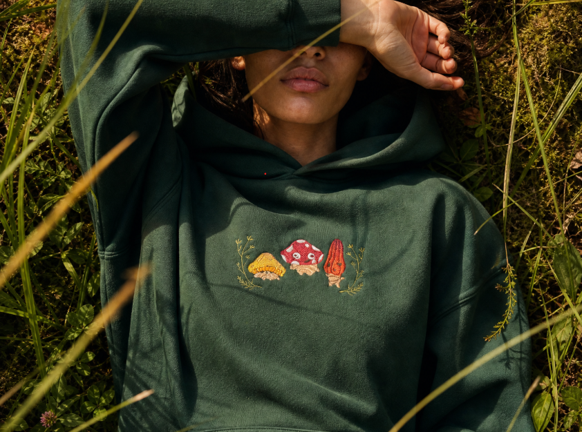 A person lying in grass outdoors wearing a dark green hoodie with a small embroidered mushroom graphic on the chest.